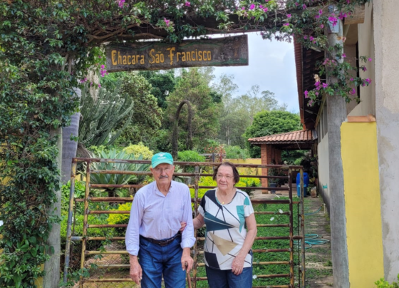 Um casal de idosos em frente a uma porteira de ferro. Sobre a cabeça há uma placa que indica o nome do sítio.