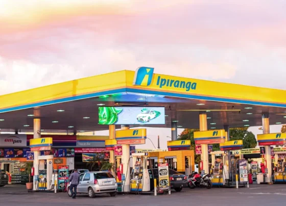 Posto de combustíveis da marca Ipiranga, com design moderno em amarelo e azul, bombas de abastecimento e uma loja de conveniência ampm, capturado ao entardecer com vegetação ao fundo.