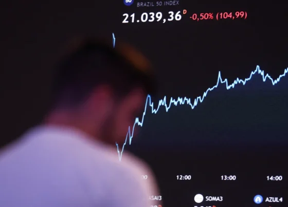 Gráfico de linhas mostrando a queda de 0,50% no índice Brazil 50, com uma figura humana desfocada em frente à tela exibindo variações de preços e logotipos de empresas na parte inferior.