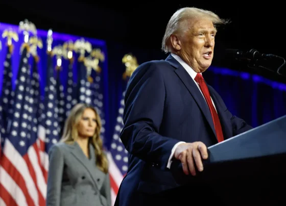 O presidente eleito dos EUA, Donald Trump, um homem em terno azul escuro discursando em um palco com bandeiras americanas ao fundo, observado por sua esposa, Melania Trump, uma mulher de casaco cinza.