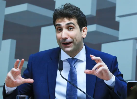 Retrato do economista Gabriel Galípolo, um homem de cabelo escuro, vestindo um terno azul escuro, camisa branca e gravata azul clara, gesticulando enquanto fala ao microfone com um fundo de figuras geométricas cinza.