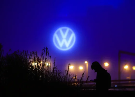 Uma pessoa segura um dispositivo sob o logotipo iluminado da Volkswagen em um cenário noturno com plantas e luzes ao fundo.