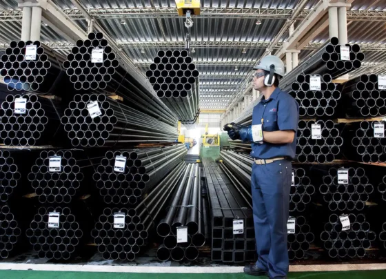 Imagem de um armazém industrial com pilhas de tubos metálicos e um trabalhador de uniforme azul, equipado com equipamentos de segurança, segurando um dispositivo.